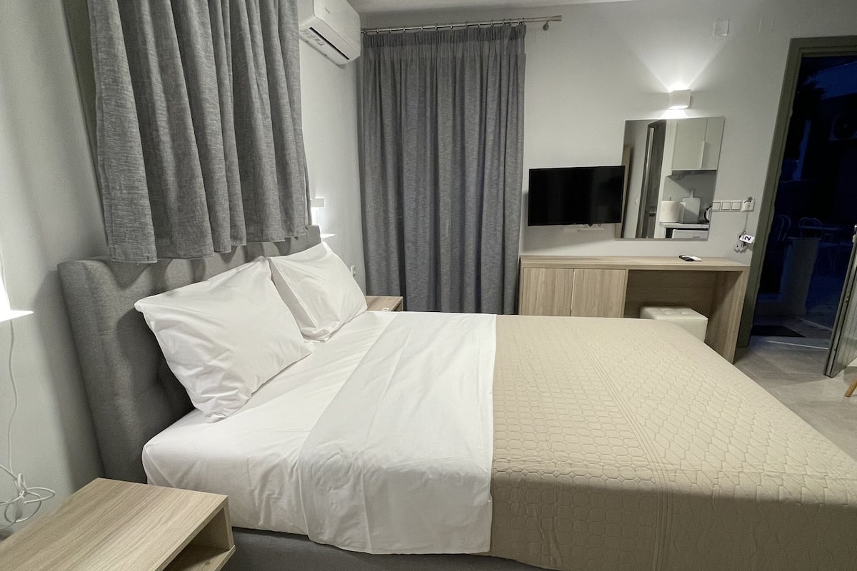 A modern hotel room with a large bed, a bedside table, a desk, a flat-screen TV, and a mirror.