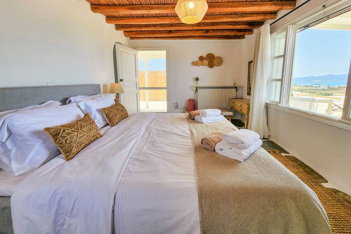 Ein Hotelzimmer mit einem großen Bett, Holzboden, Blick aufs Meer und einem kleinen Tisch mit einer Lampe.