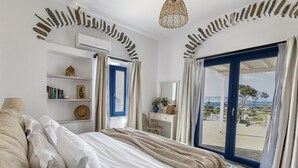 A bedroom with a bed, a desk, a chair, a shelf with books, a hat, a vase with flowers, and a view of the sea through a door. - Anthemion Paros Villas & Suites (Paros)
