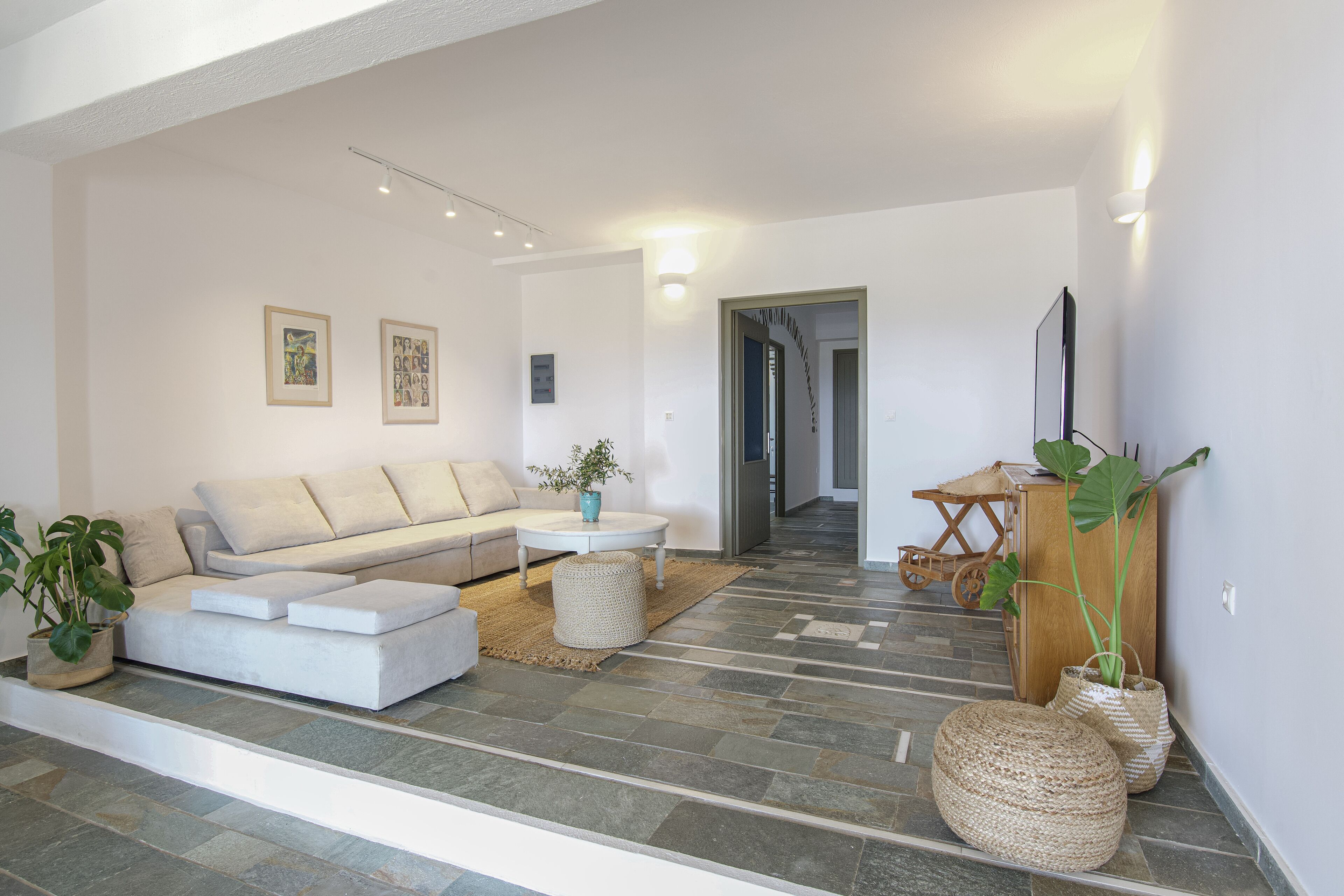 Ein modernes Wohnzimmer mit einem Ecksofa, einem Couchtisch und einem Fernseher. Zwei Zimmerpflanzen und ein gewebter Hocker ergänzen das Bild. Der Raum präsentiert eine neutrale Farbgestaltung und einen Steinboden.