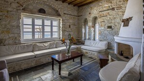 A stone-walled room with a wooden ceiling, a white sofa, a wooden table with a vase, and a fireplace. - Anthemion Paros Villas & Suites (Paros)
