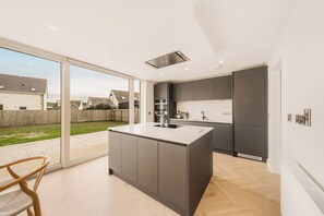 Une cuisine moderne dotée d’un grand îlot central, d’appareils électroménagers intégrés et d’une porte-fenêtre coulissante donnant sur un jardin.