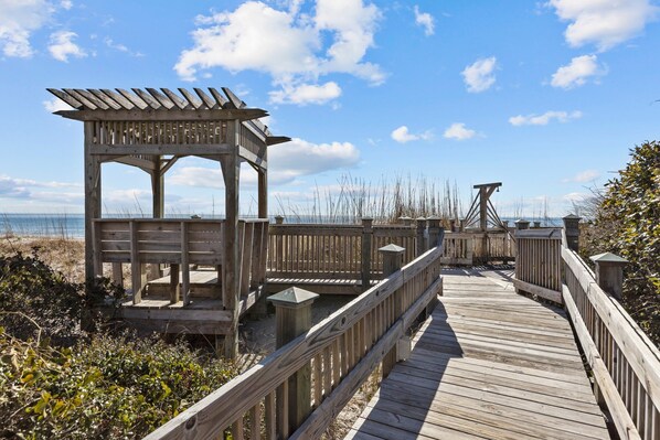 A wooden boardwalk leading to a wooden gazebo with a sloped roof, surrounded by beach vegetation and a clear sky.