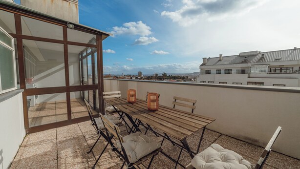 Una terraza en la azotea con mesa, sillas y cojines, con vistas a un paisaje urbano.