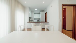 Un comedor moderno con una mesa y sillas blancas, una cocina al fondo y una puerta de madera a la derecha.
