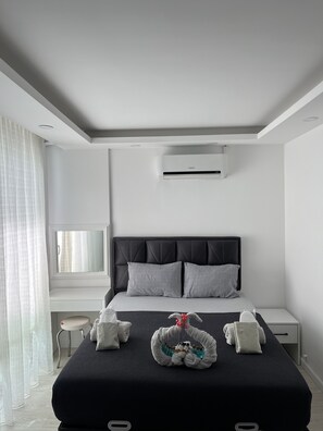 Una habitación de hotel moderna con una cama grande, una mesita de noche, un escritorio pequeño y una ventana con cortinas traslúcidas.
