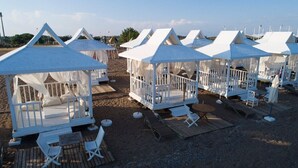 Strandhütten mit Betten und Sitzgelegenheiten, auf einer Sandfläche mit einer Holzterrasse angeordnet.