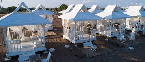 Beach huts with beds and seating, arranged on a sandy area with a wooden deck.