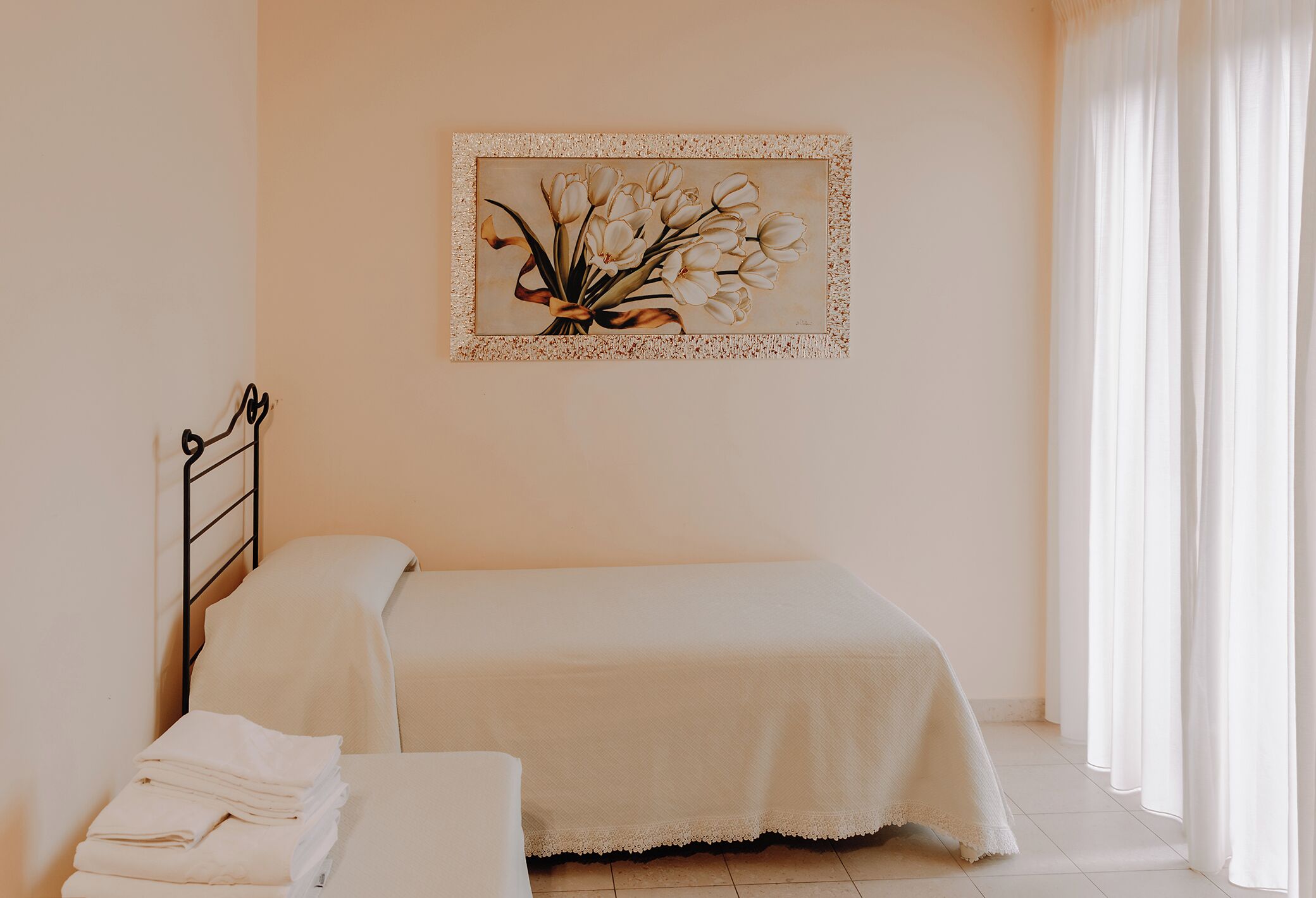 A neatly made bed with a white bedspread, a small bedside table with folded towels, and a framed floral artwork on the wall.