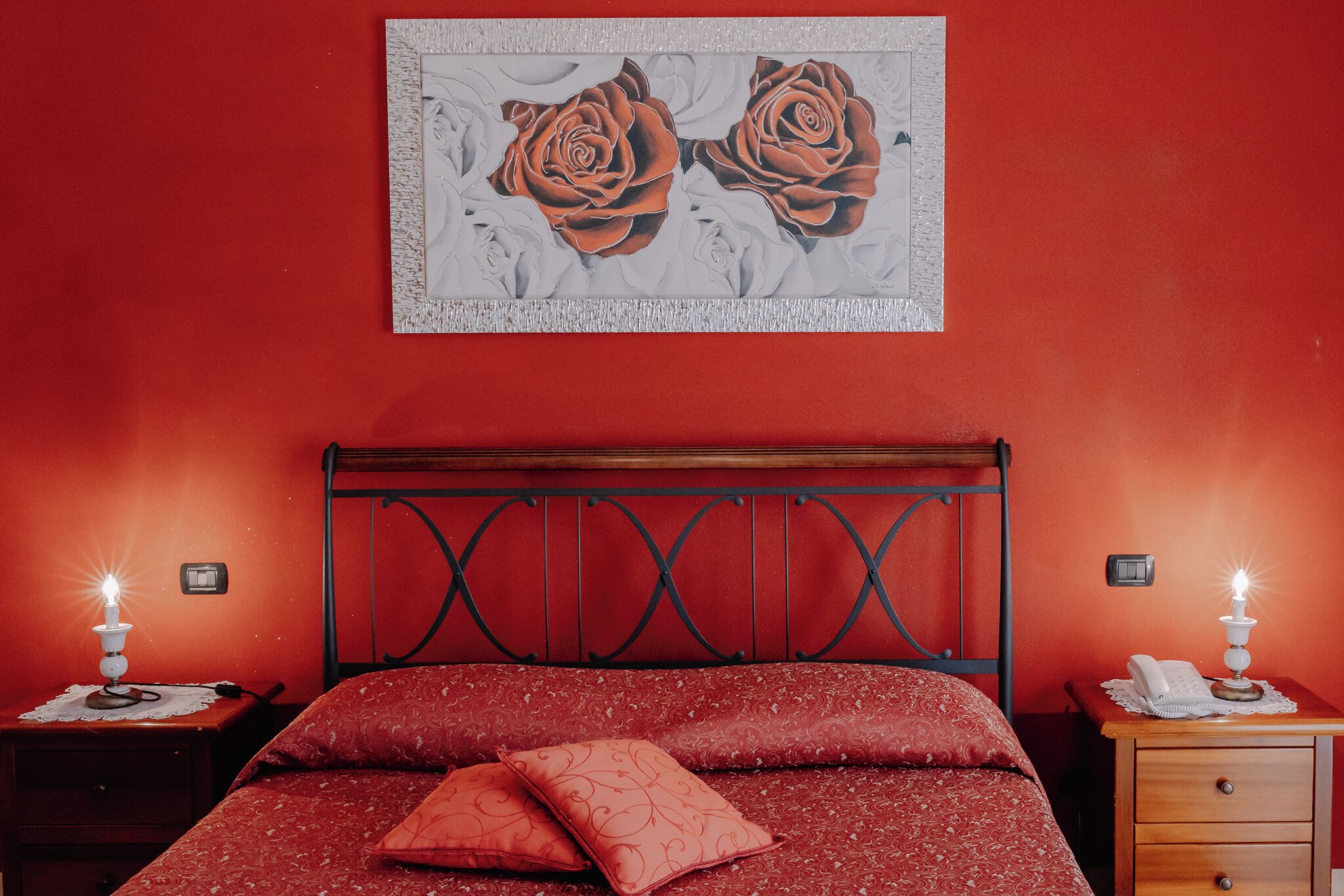 Un dormitorio con una pared roja, una cama con ropa de cama roja, dos mesitas de noche con lámparas y un cuadro de rosas sobre la cama.