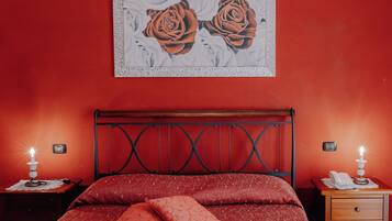 Un dormitorio con una pared roja, una cama con ropa de cama roja, dos mesitas de noche con lámparas y un cuadro de rosas sobre la cama.