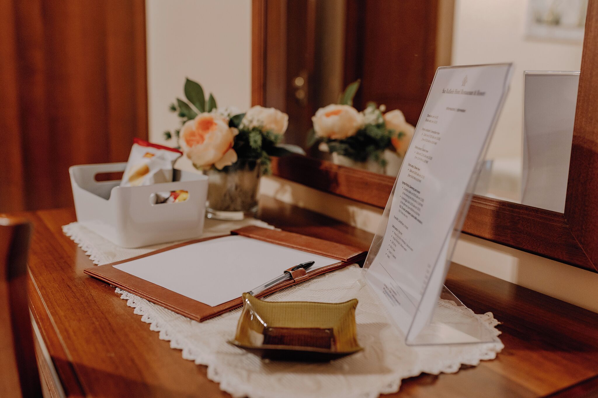 Una scrivania in legno con un sottobicchiere in pizzo bianco, un blocchetto per appunti, una penna, una piccola borsa gialla, una scatola di fazzoletti bianchi e un espositore per menu.