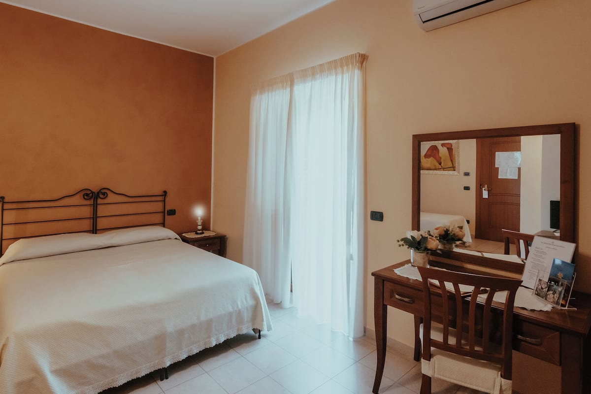 Un dormitorio con una cama, un escritorio con espejo, una mesita de noche y una ventana con cortinas.