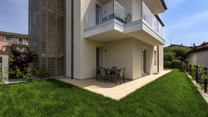 A modern two-story house with a balcony, a patio with a table and chairs, and a well-maintained lawn.
