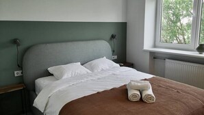 Una cama bien hecha con sábanas blancas y una manta marrón, una mesita de noche con una lámpara, y una ventana con vistas a árboles.