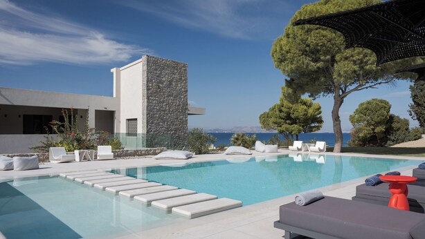A modern pool area with lounge chairs and a stone wall, overlooking a scenic view. - Villa Vel Aegina (Aegina)
