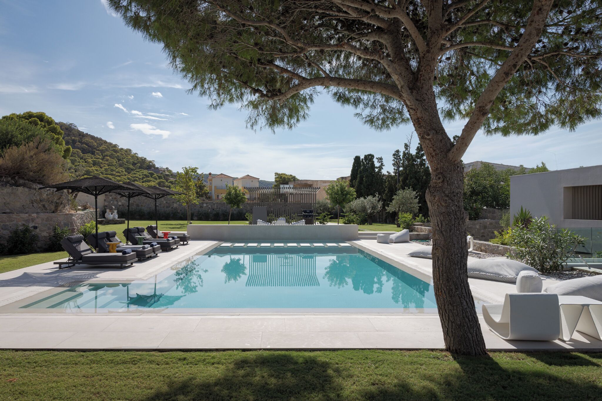 A rectangular swimming pool with a clear blue water surface, surrounded by a low white tiled deck. There are several lounge chairs and a small covered seating area with a table. A large tree with a thick trunk and dense foliage is prominently in the foreground.