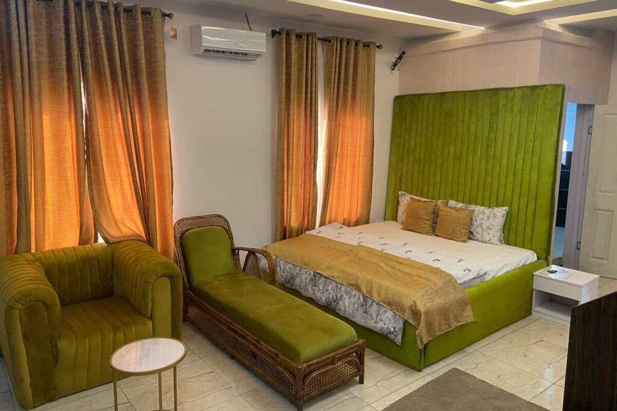 Un dormitorio con una cama verde, un sillón verde, una silla de mimbre y una pequeña mesa redonda.