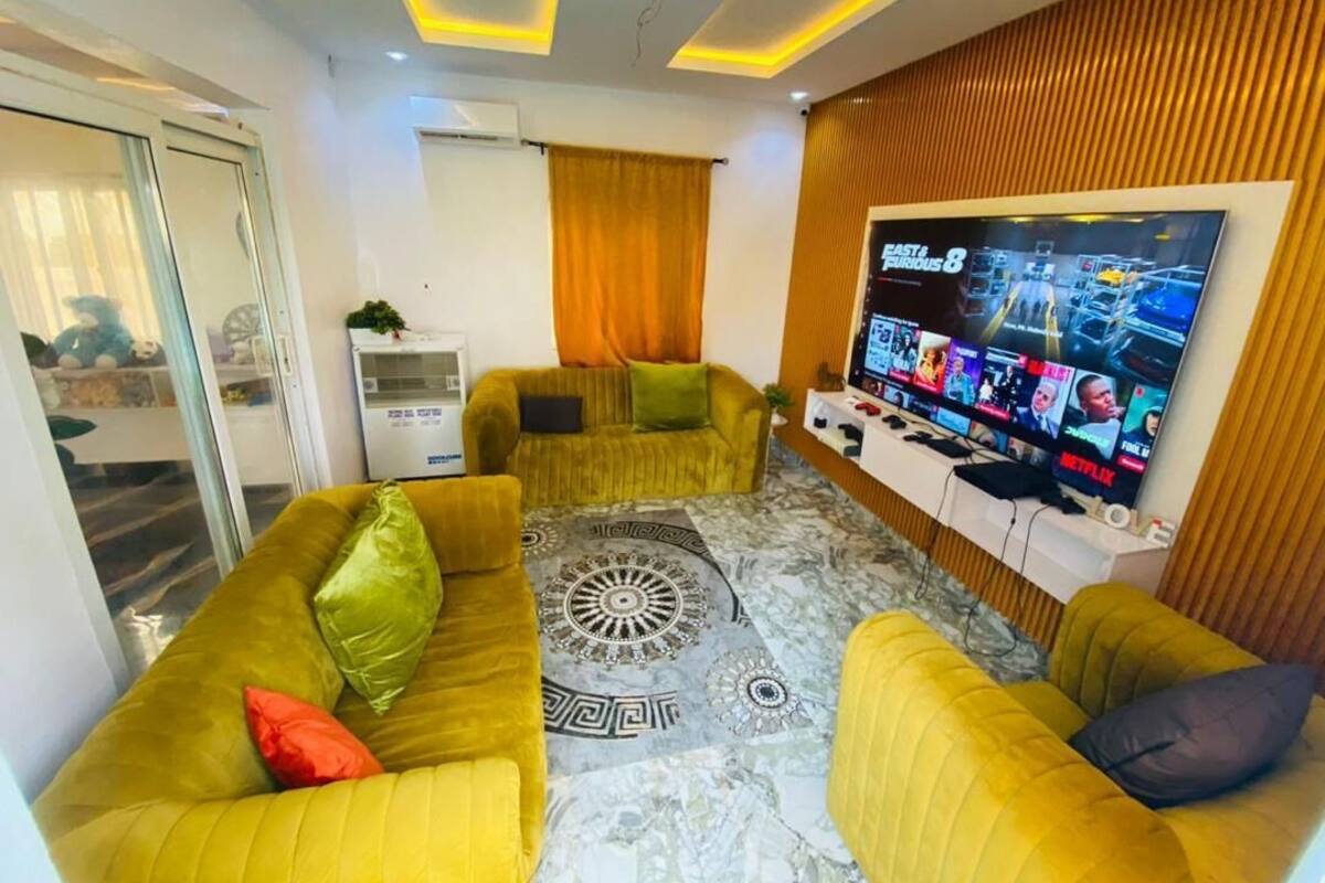 Una sala de estar con un televisor de pantalla plana grande, sofás amarillos, un tapete estampado y un panel de pared de madera.