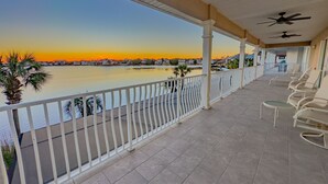 A covered balcony with a view of a sunset over a body of water, white railings, and several chairs.