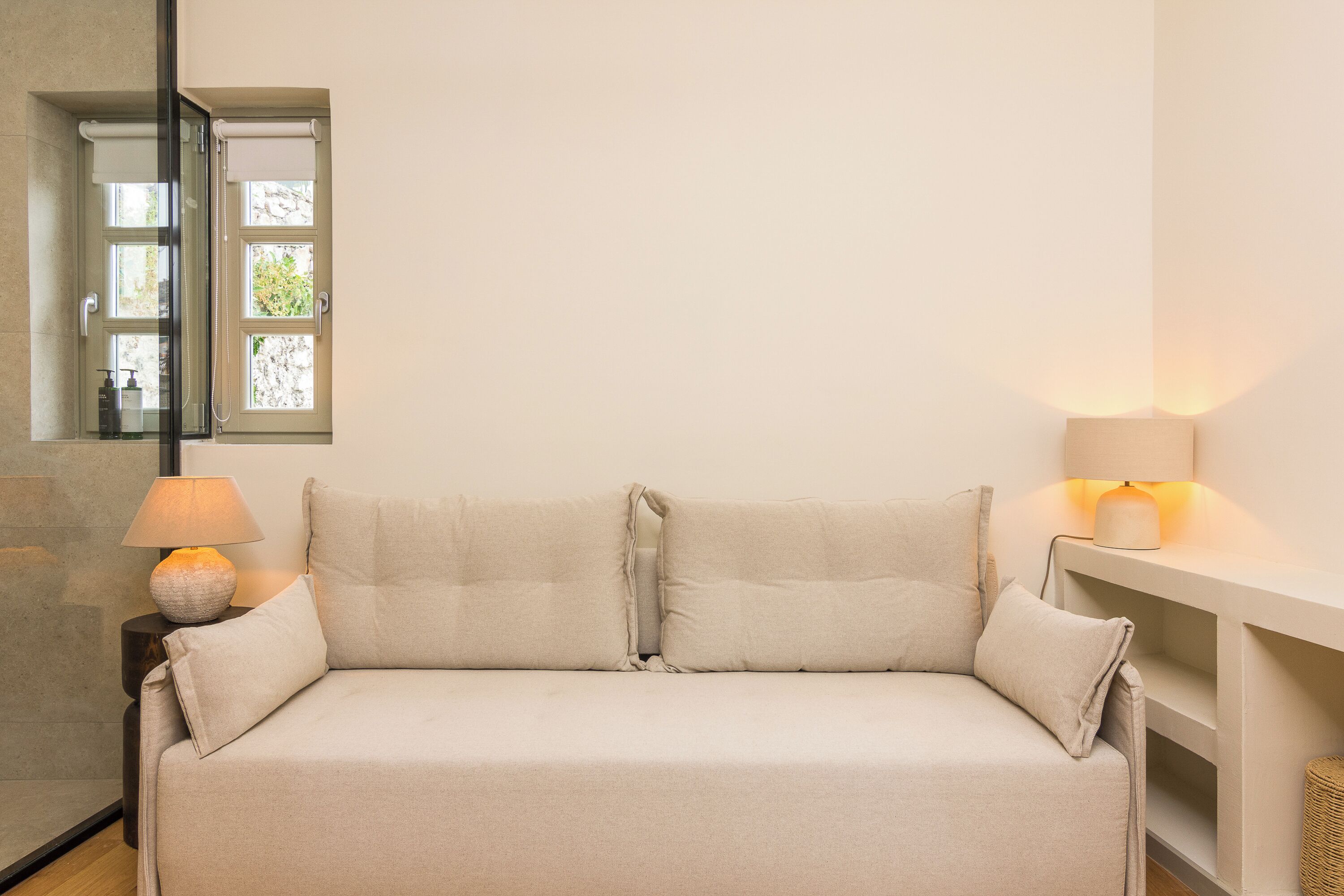 A neutral-colored sofa with two pillows, a lamp on a side table, and a window with blinds.