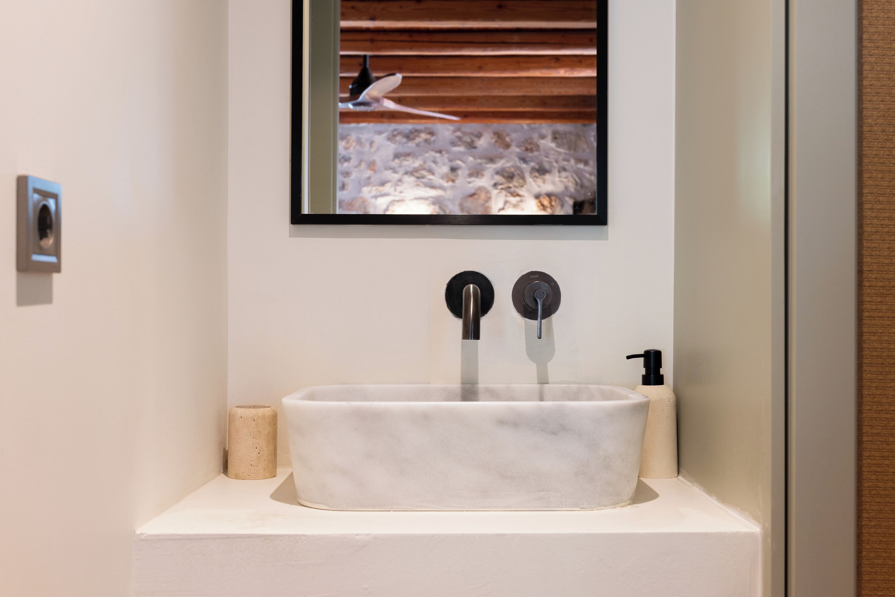 Un baño con lavamanos de mármol, dos grifos, dispensador de jabón y techo de madera.