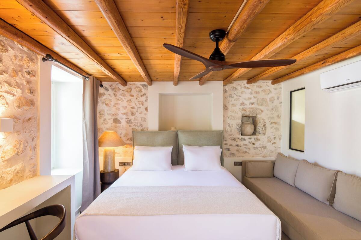 Une chambre d’hôtel avec un plafond en bois, un lit avec du linge de maison blanc, une table de chevet avec une lampe, un canapé et un mur en pierre.