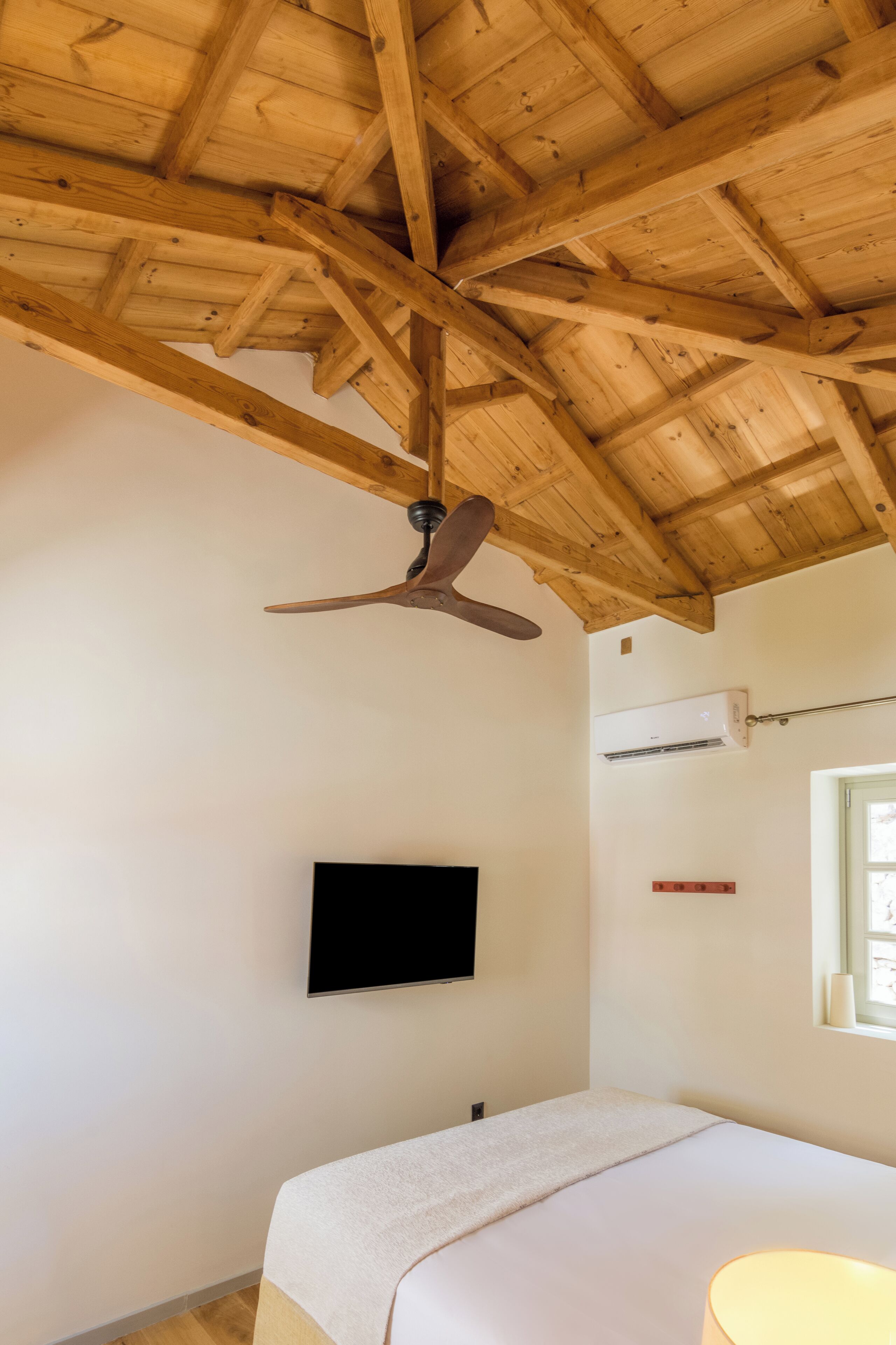 A bedroom with a wooden ceiling, a ceiling fan, a flat-screen TV mounted on the wall, and a window with a view.