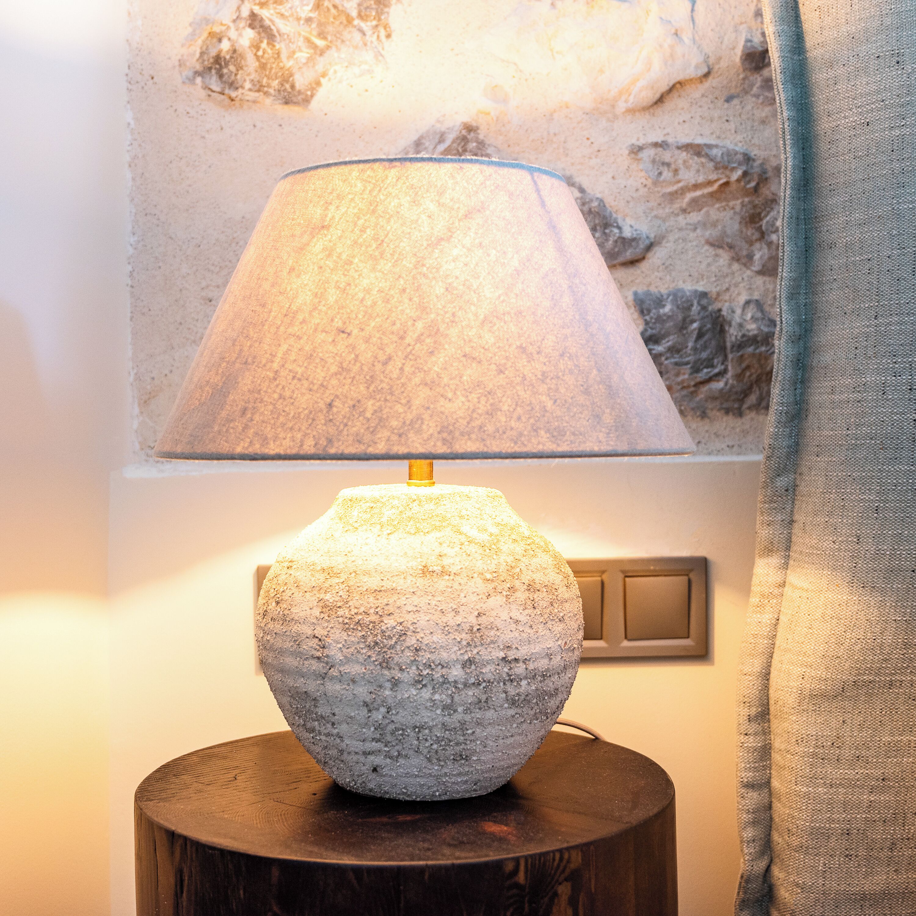 A stone lamp on a wooden table with a wall-mounted light switch and a stone wall in the background.