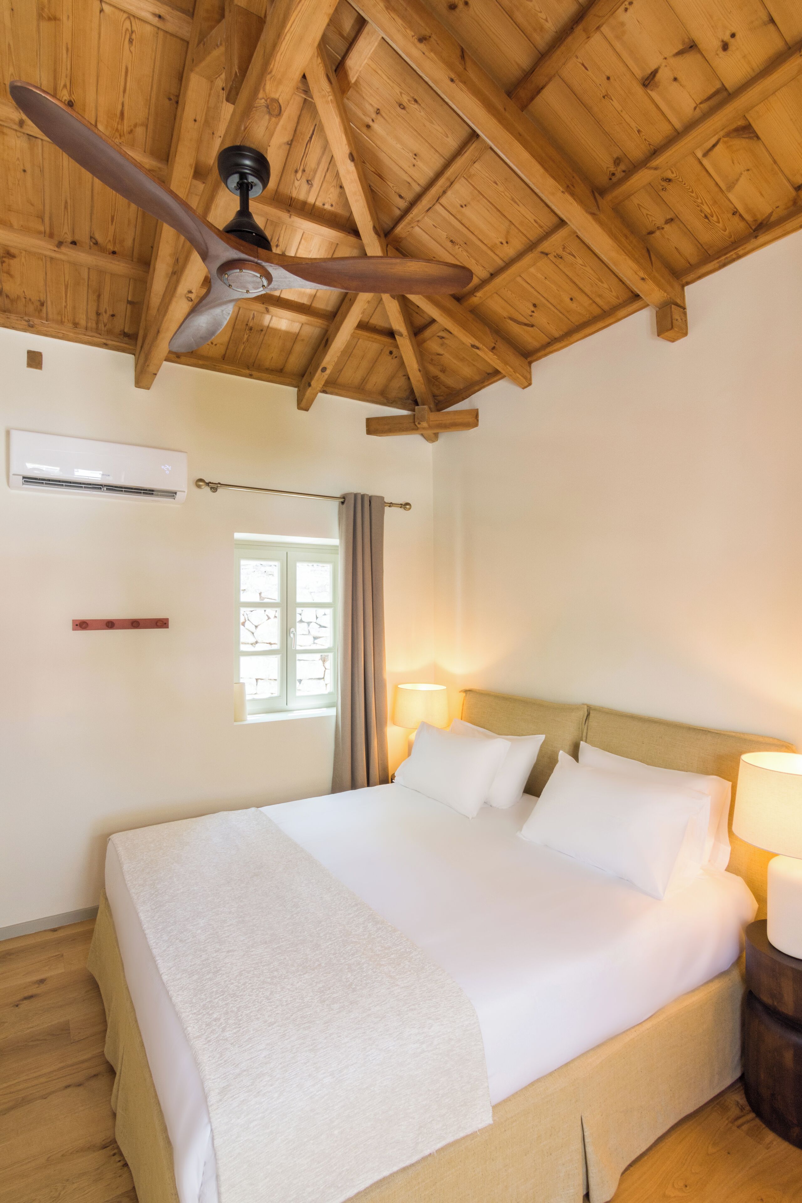 A bedroom with a wooden ceiling, a bed with white linens, a ceiling fan, a window with curtains, and a bedside lamp.