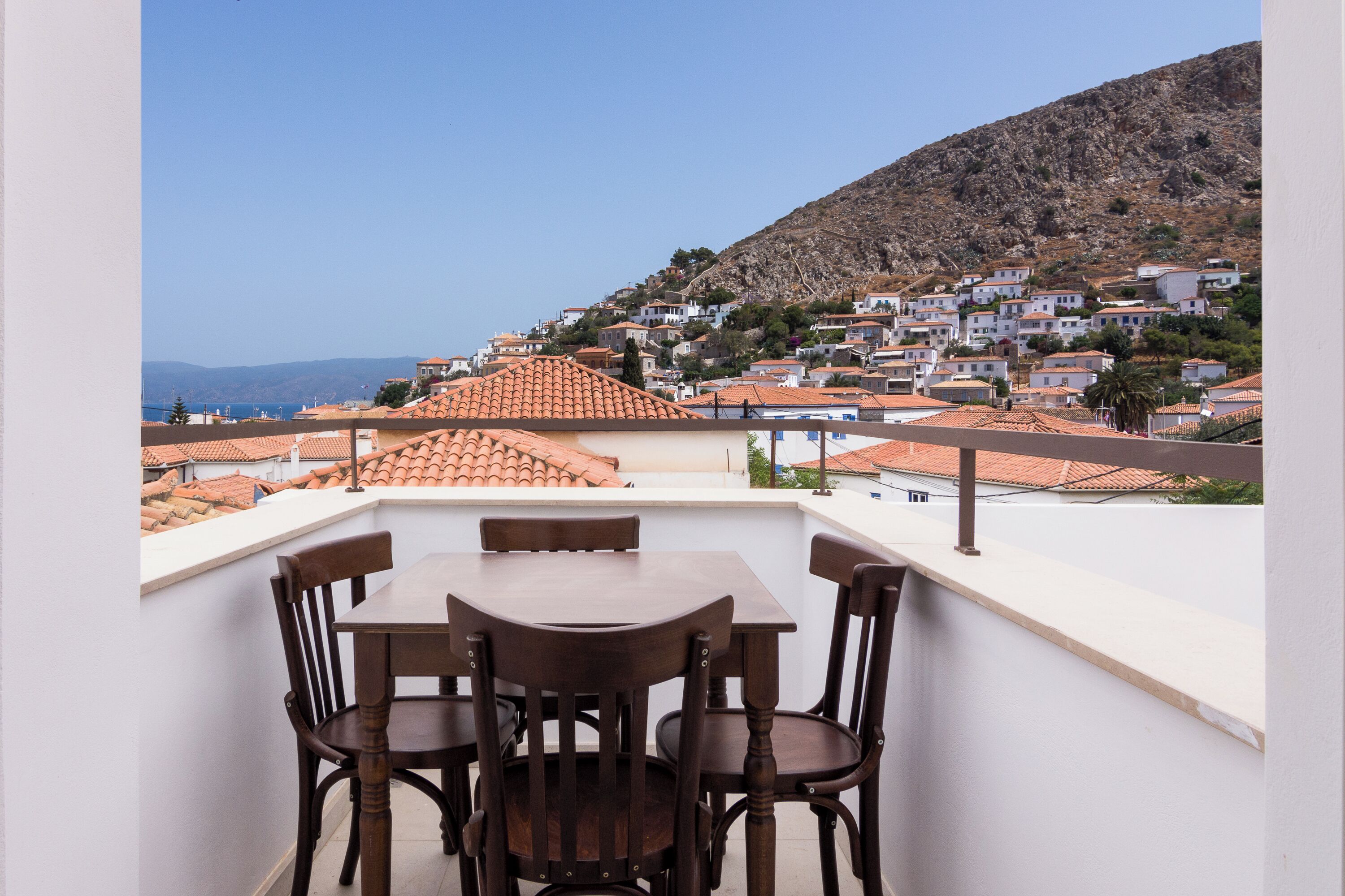 Un balcón con mesa y sillas, con vista a una colina y edificios con tejados de tejas.