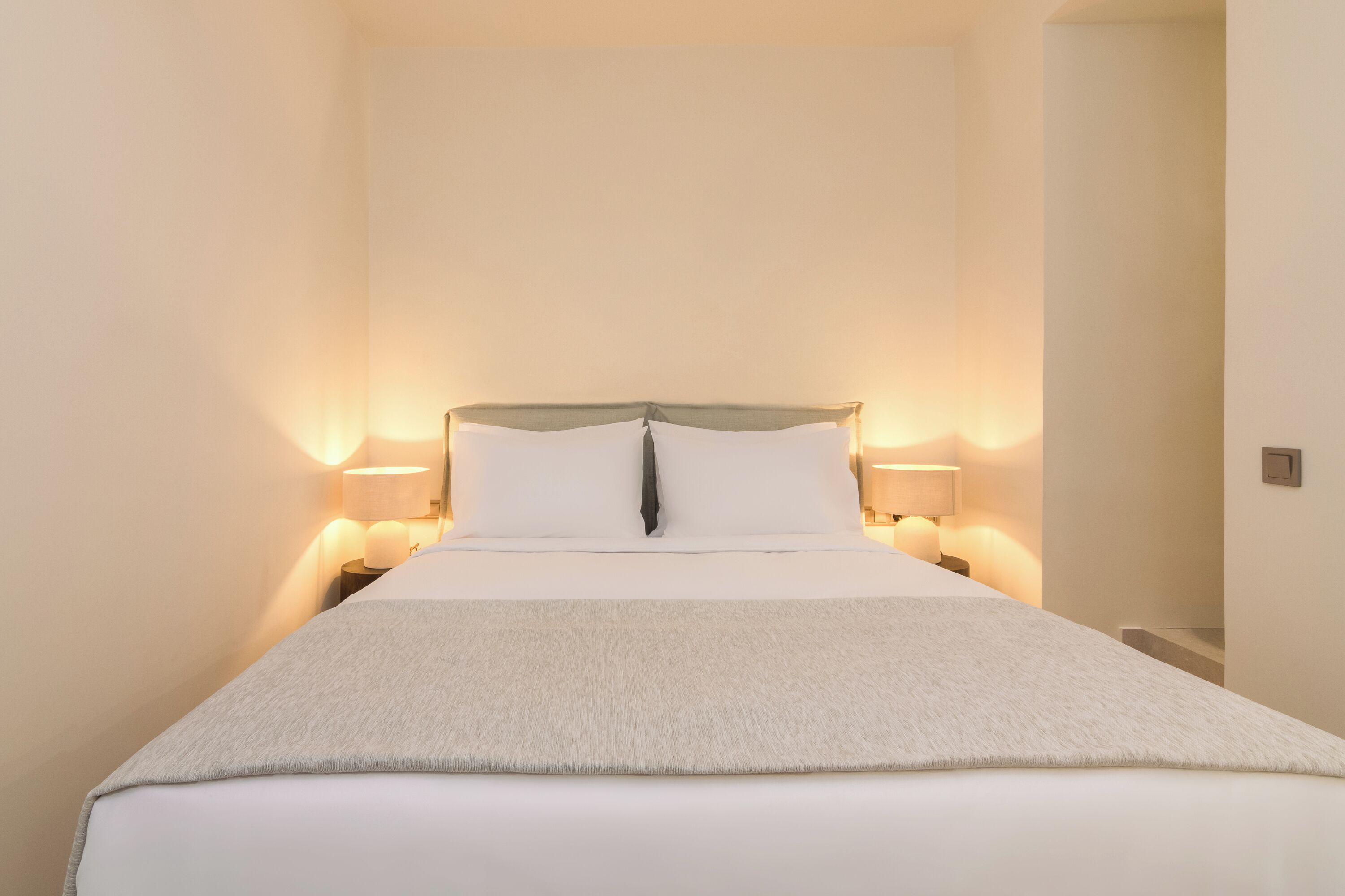 A neatly made bed with white linens and a grey throw blanket, flanked by two bedside lamps on either side.