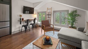Un espace de vie chaleureux de style loft, avec un plancher en bois, un canapé confortable, une petite table avec une bougie, un téléviseur fixé au mur et une grande fenêtre donnant sur un paysage verdoyant.