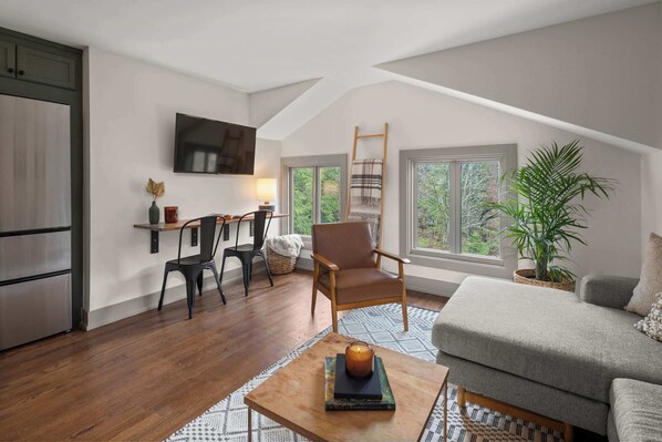 A cozy loft-style living space with a wooden floor, a comfortable sofa, a small table with a candle, a TV mounted on the wall, and a large window offering a view of greenery outside. - Stowe Triple Condo | 6BR Mountain & River Views (Stowe)