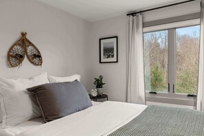Quarto com cama, almofadas, mesa de cabeceira com planta e relógio, quadro emoldurado e janela com cortinas.