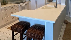 A modern kitchen with white cabinets, a stainless steel refrigerator, a microwave, and a sink. There are two woven stools at the island. - Elegant condo with beach views - Sunset Royale 314 (Siesta Key)