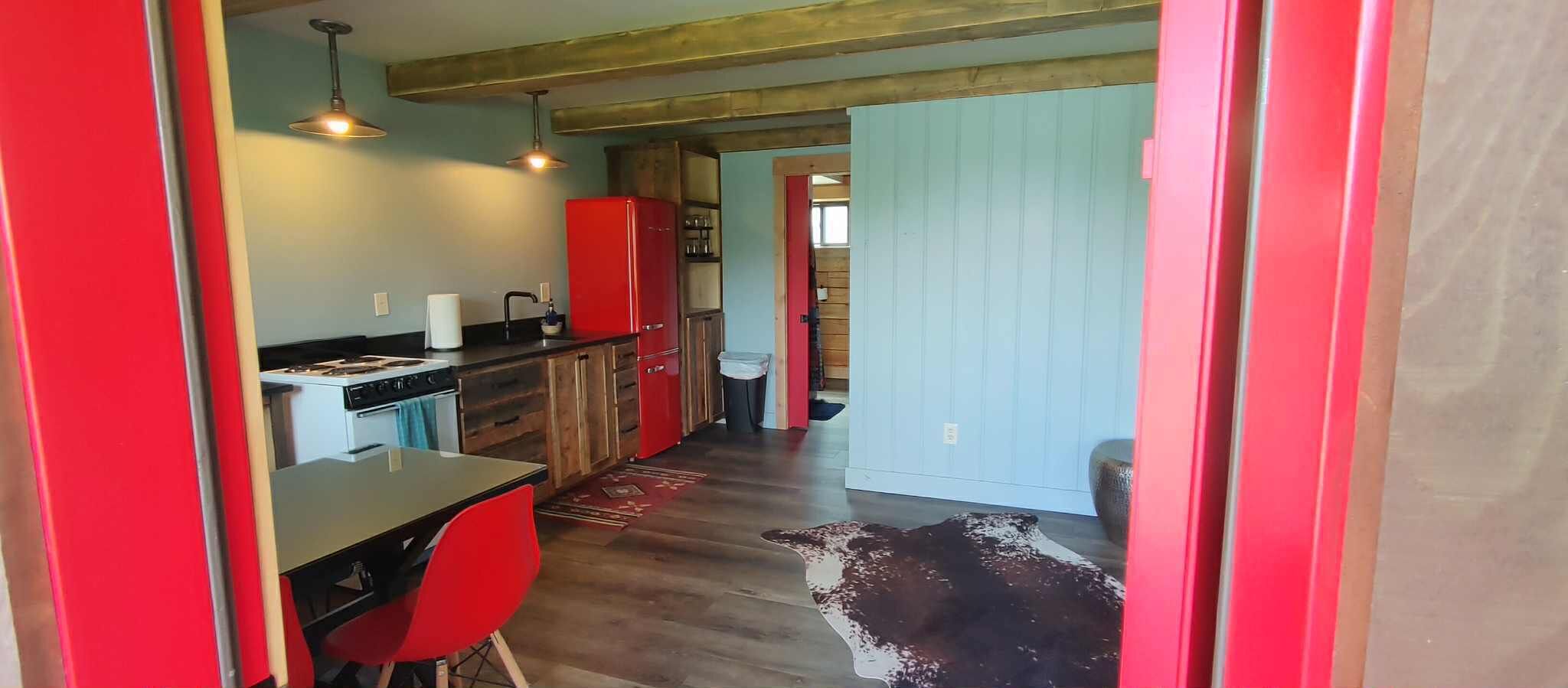 A compact kitchen with a stove, sink, and red cabinets. A dining table with red chairs is positioned in front of the kitchen.