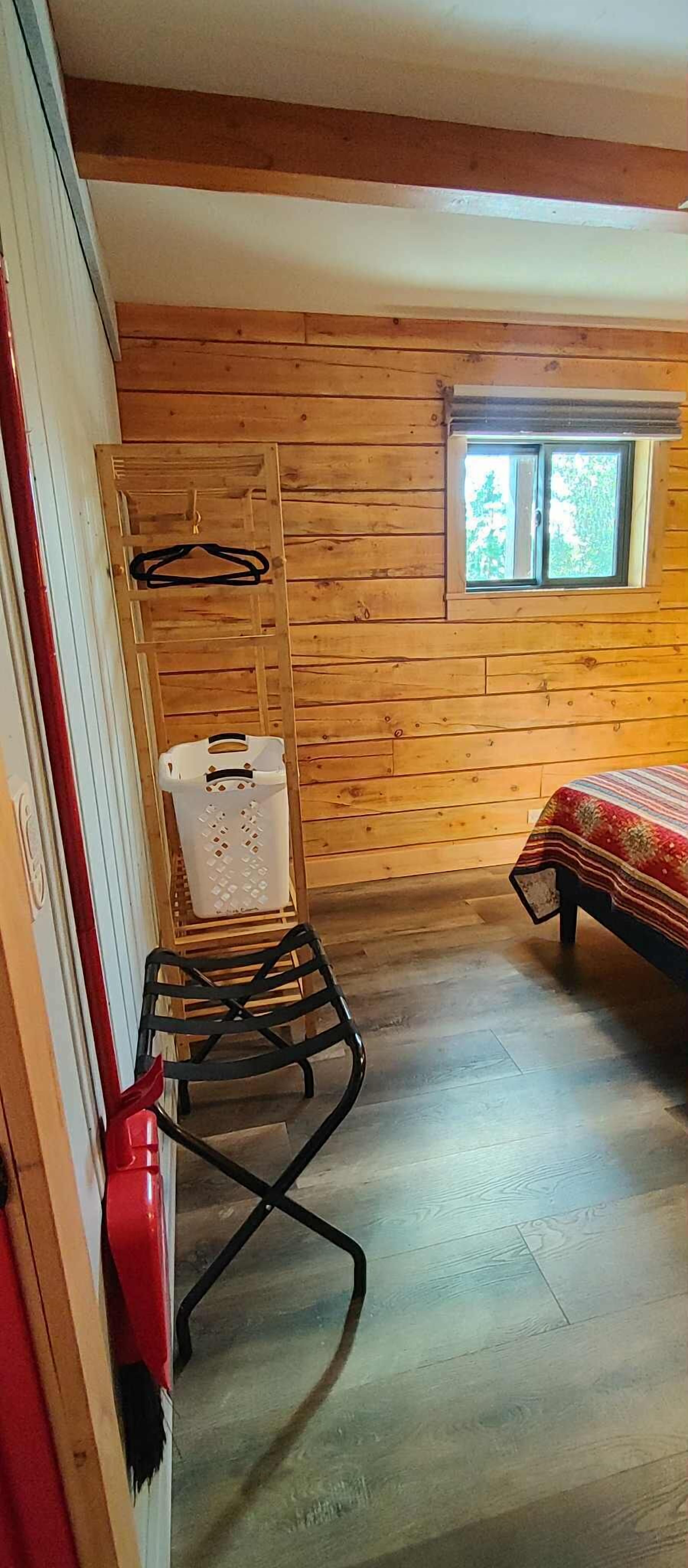 A wooden cabin room with a bed, a window, a drying rack, and a red fire extinguisher.