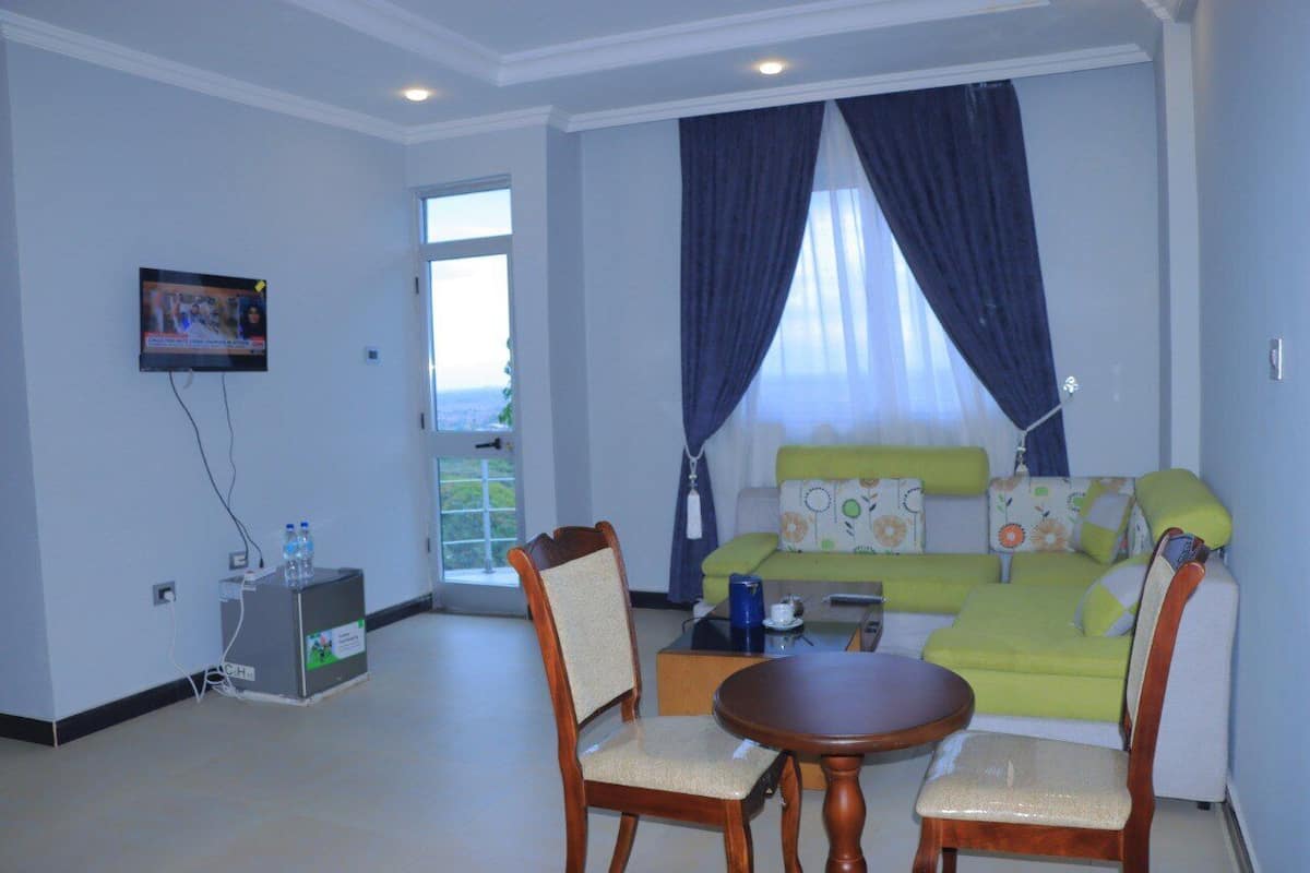 A living room with a green sofa, wooden chairs, a small table, a TV, and a window with curtains.