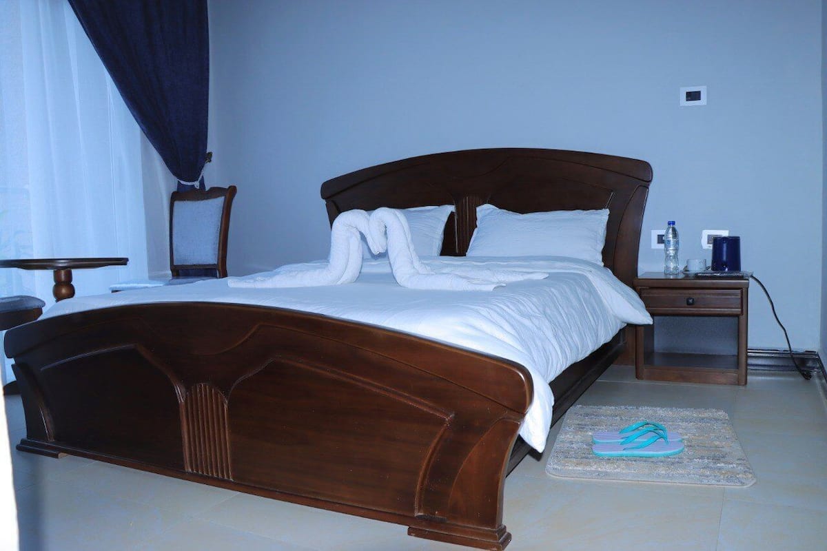 A neatly made bed with a wooden headboard, a small bedside table with a lamp, and a pair of slippers on a mat.