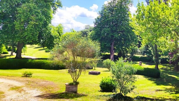 Un jardin bien entretenu, avec différents arbres et arbustes, un ciel dégagé et un chemin de terre.