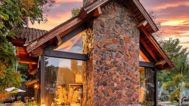 A stone-clad building with a large glass window and a patio area with outdoor furniture. - LAS HOTEL BOUTIQUE (Foz do Iguaçu)