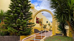 A yellow building with a staircase leading to an entrance, surrounded by greenery and a flower bed.