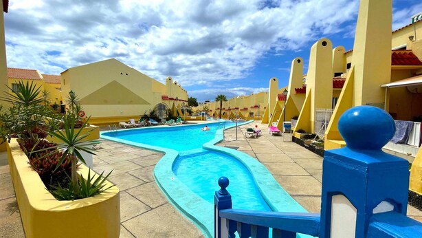 A swimming pool with a curved design, surrounded by yellow buildings with red-tiled roofs, and a clear blue sky.