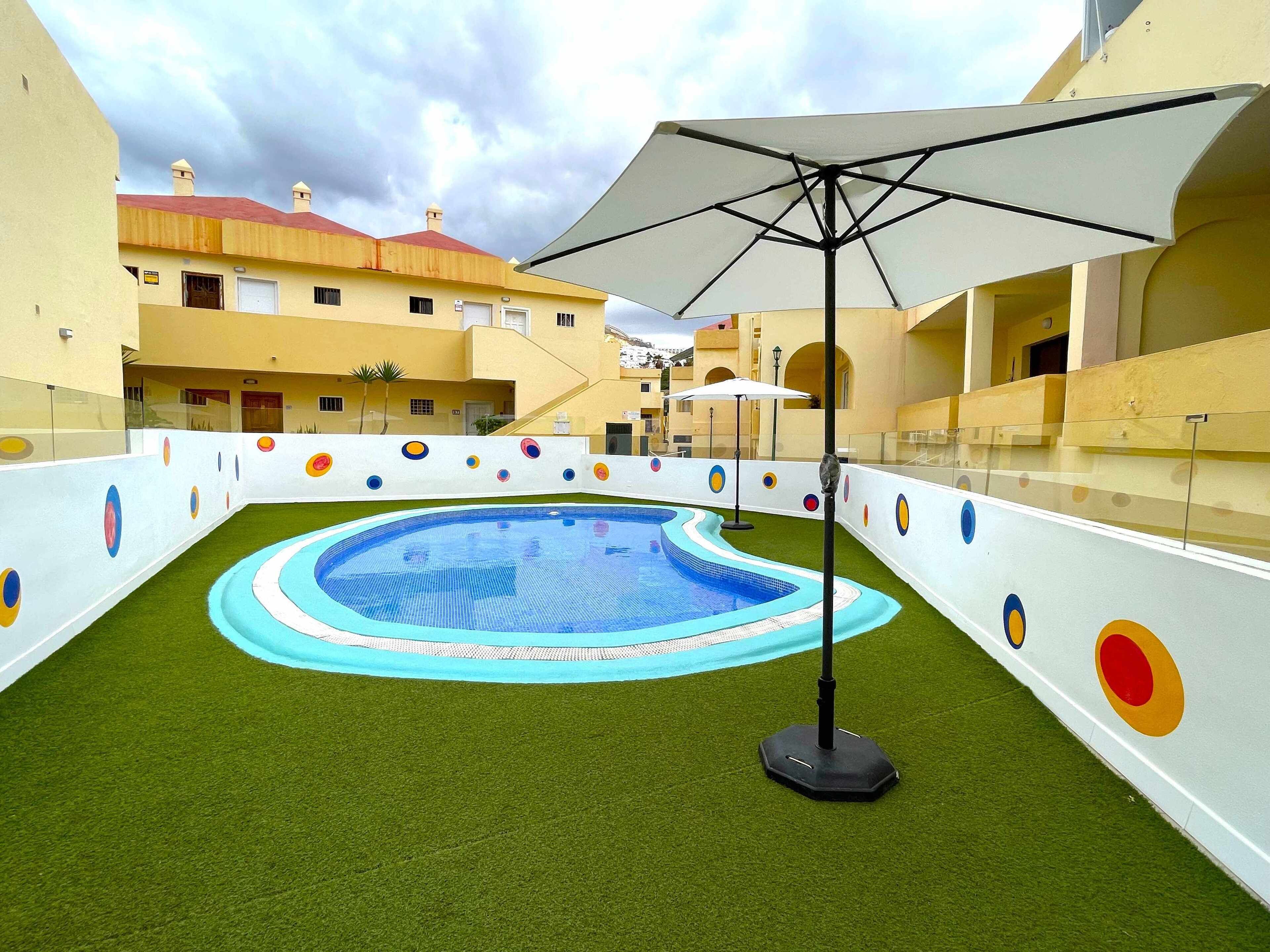 Une piscine entourée d’une terrasse, un espace de repos couvert et, au fond, un bâtiment avec un toit rouge.