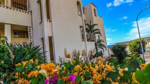 Un immeuble à plusieurs étages doté de balcons, un jardin parsemé de fleurs jaunes et un ciel d’un bleu limpide.