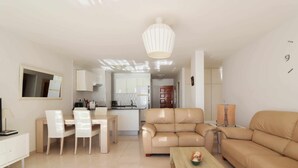 Un salon moderne avec un canapé beige, un coin repas avec des chaises blanches et une cuisine en arrière-plan.