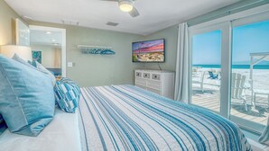 A bedroom with a bed, a ceiling fan, a TV, and a view of the beach.