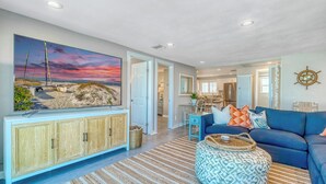A living room with a large TV, a blue sofa, patterned ottoman, and a striped rug.