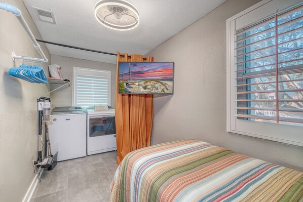 Un dormitorio con cama, ventana con persianas, televisor en la pared, lavadora y ventilador de techo.